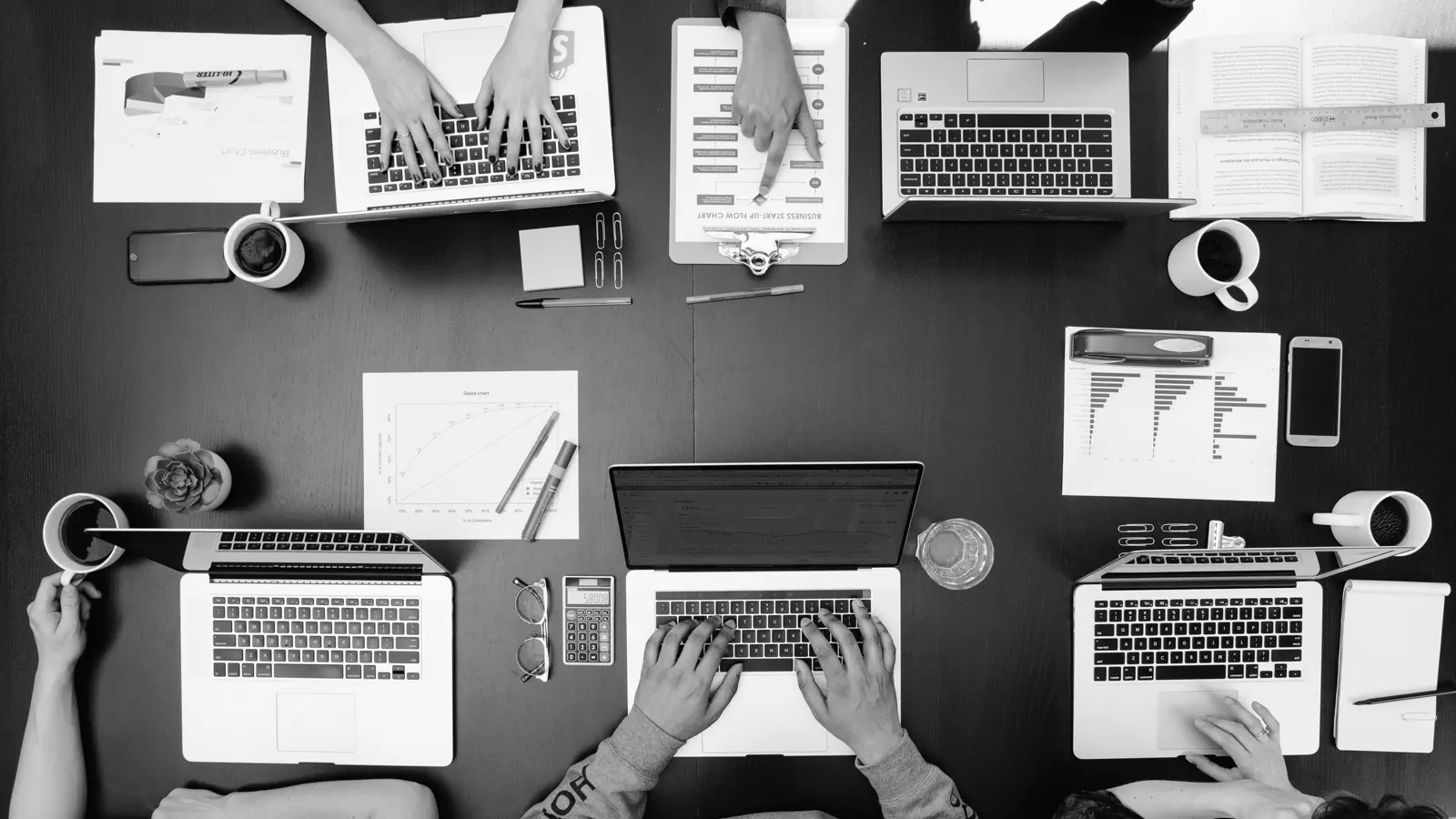 Vue de dessus d'une table de travail : ordinateurs portables, graphiques imprimés, carnets et cafés.